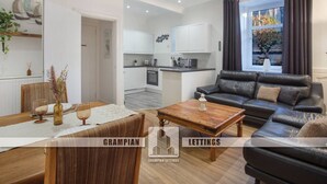 A modern living room with a black leather sofa, wooden coffee table, and a dining area with a table set for two. A kitchen is visible in the background. - Selkies Retreat | Grampian Lettings Ltd (Aberdeen)