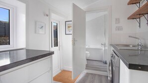 A modern kitchen with white cabinetry, a black countertop, and a built-in dishwasher. There is a window on the left side and a door leading to a bathroom with a white toilet and bathtub. - Selkies Retreat | Grampian Lettings Ltd (Aberdeen)