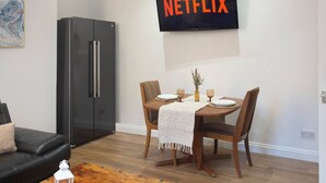 A modern living room with a dining area, a black refrigerator, a flat-screen TV mounted above a wooden dining table, and a black couch. - Selkies Retreat | Grampian Lettings Ltd (Aberdeen)