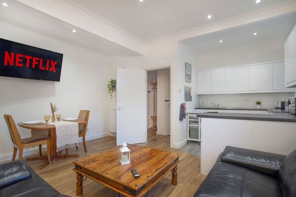 A modern living room with a wooden coffee table, a leather sofa, a dining area with a white table and chairs, a kitchen with white cabinets, and a TV mounted on the wall displaying 'NETFLIX'.