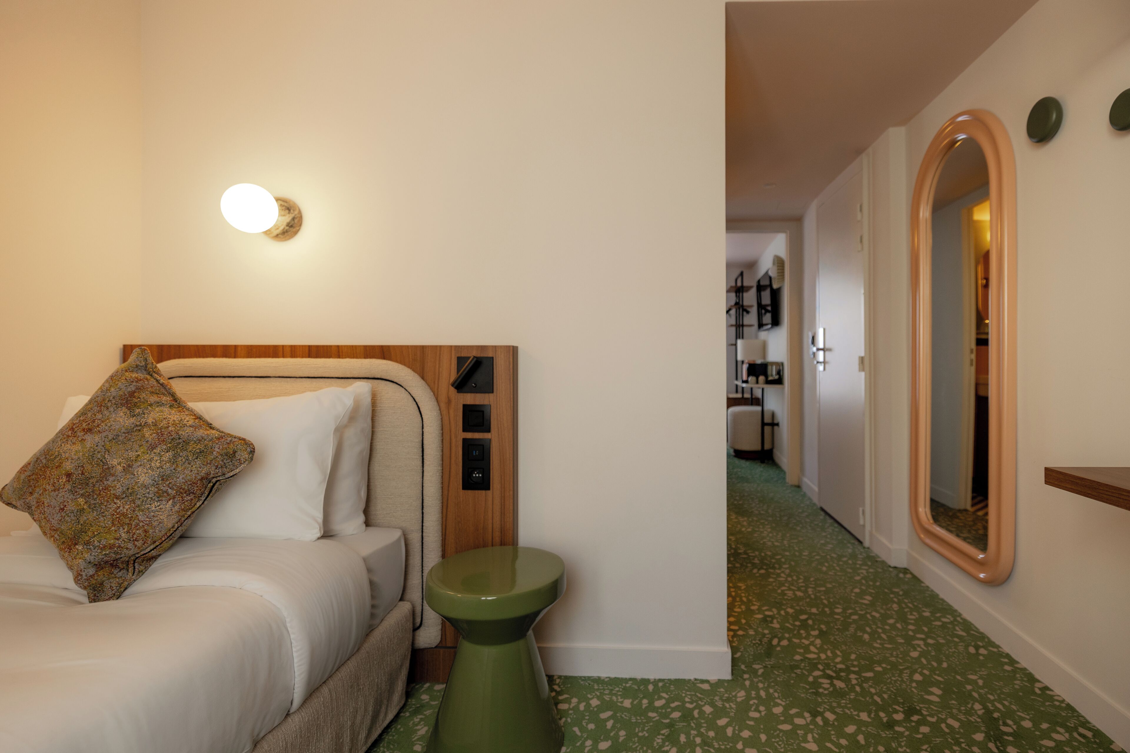 A hotel room with a bed, a small stool, a wall-mounted mirror, and a hallway leading to other rooms.