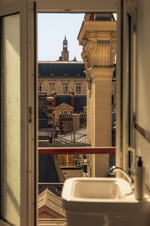 Une vue depuis une fenêtre d’un bâtiment, avec un grand évier blanc, une rambarde en métal et un aperçu d’un paysage urbain caractérisé par une architecture historique.