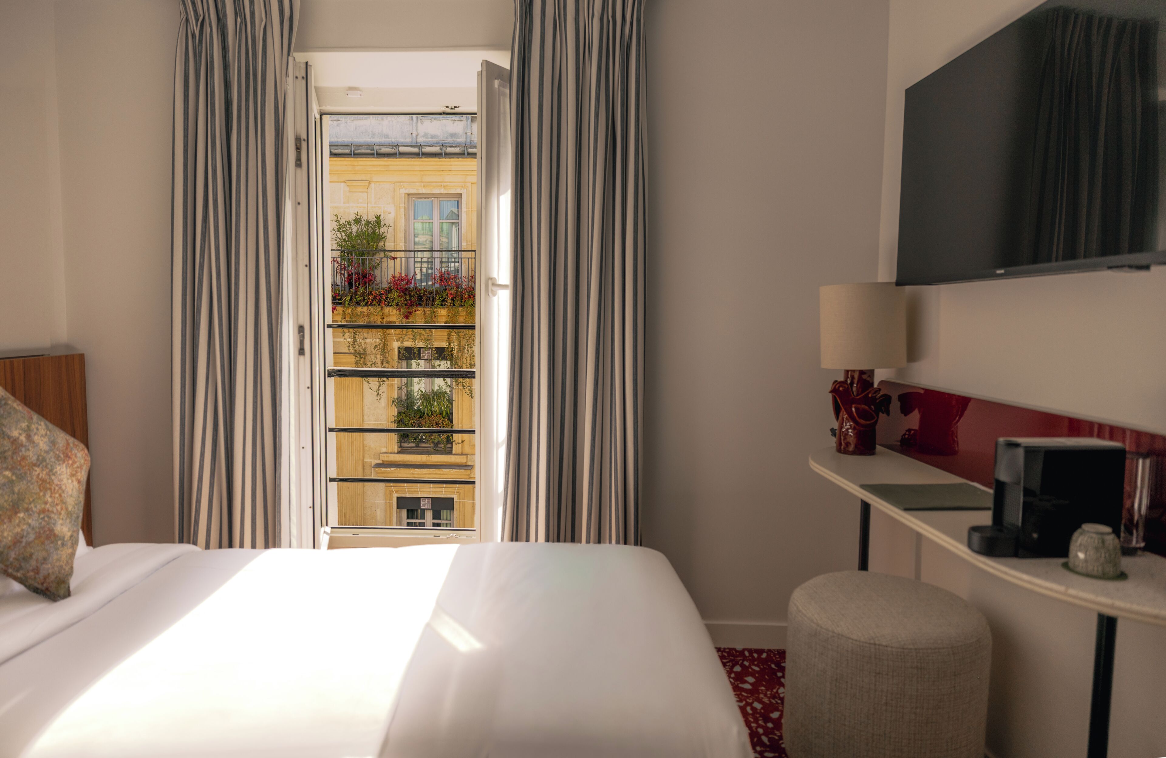 A hotel room with a bed, a desk, a TV, and a view of a balcony with plants.