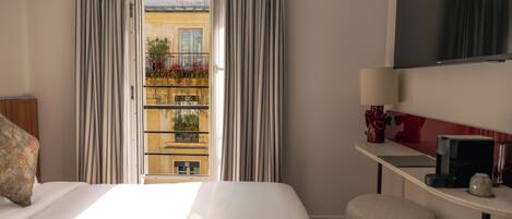 A hotel room with a bed, a desk, a TV, and a view of a balcony with plants.