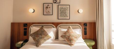 Habitación de hotel con cama, dos almohadas, una mesita de noche, un taburete verde y luces fijadas a la pared.