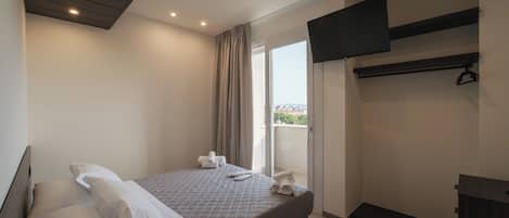 A modern hotel room with a bed, a flat-screen TV mounted on the wall, a balcony with a view, and a small desk area.