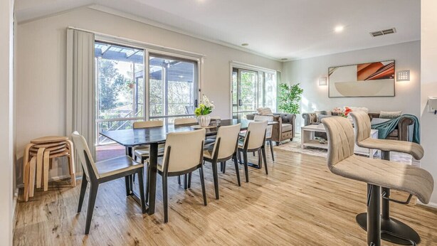Amplia sala de estar con una gran mesa de comedor, múltiples sillas, un sofá y una mesa de centro. Amplias ventanas ofrecen luz natural y vistas al exterior.