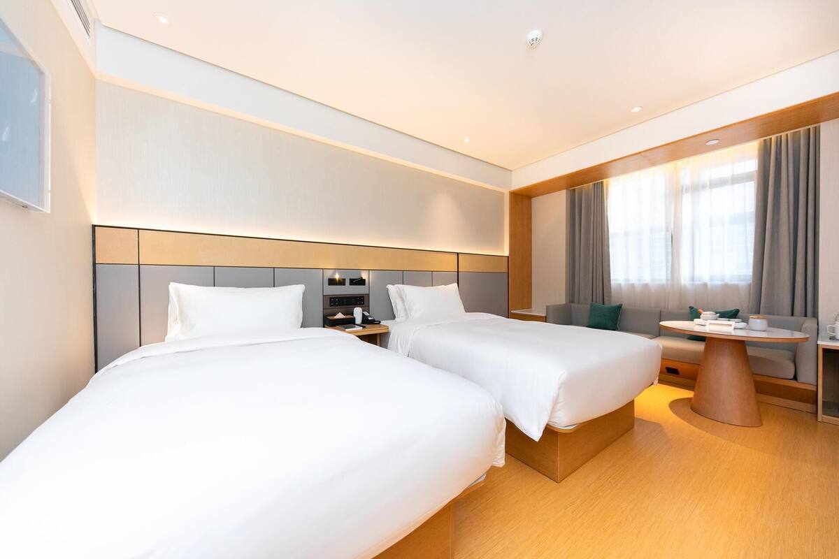 A hotel room with two beds, a small table, a sofa, and a large window with curtains.