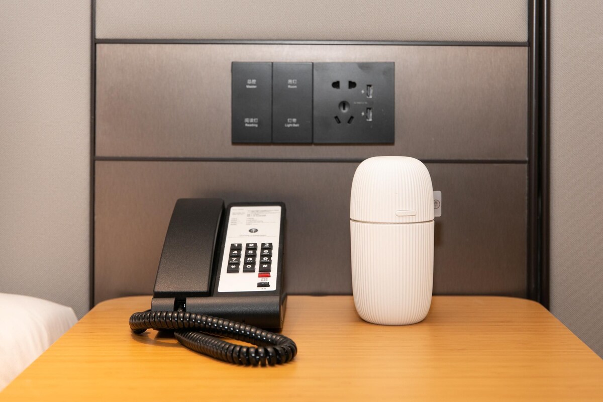 A hotel room with a telephone, a power outlet, and a water bottle on a wooden nightstand.