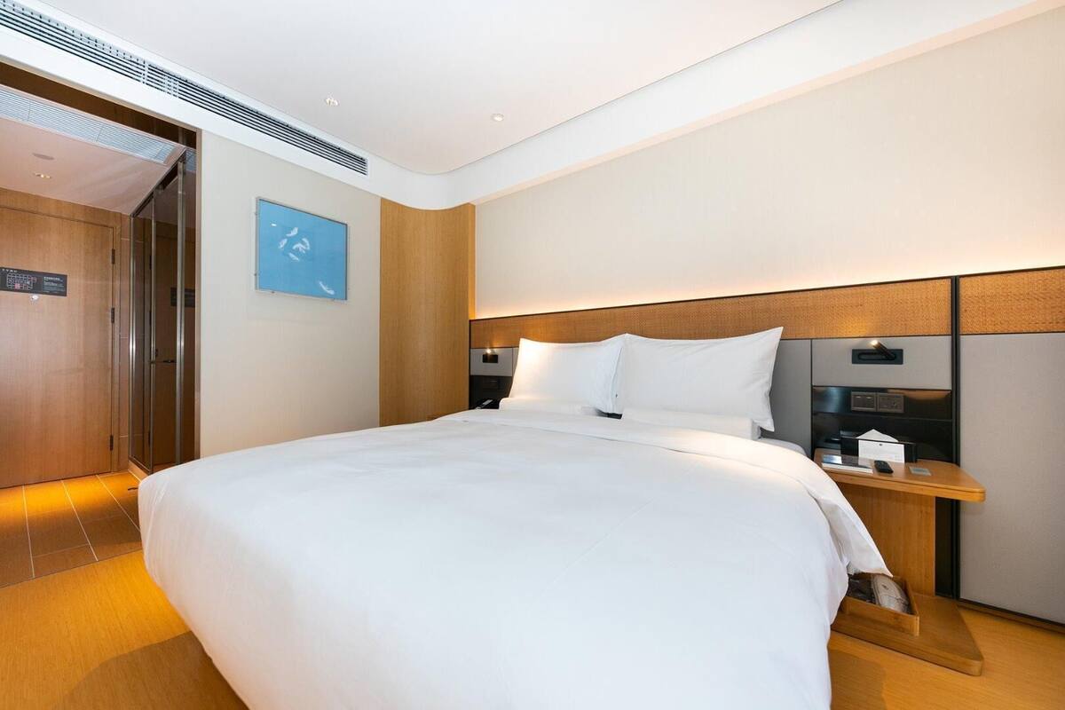 A modern hotel room with a large bed, a wooden headboard, a bedside table with a lamp, and a wall-mounted TV.