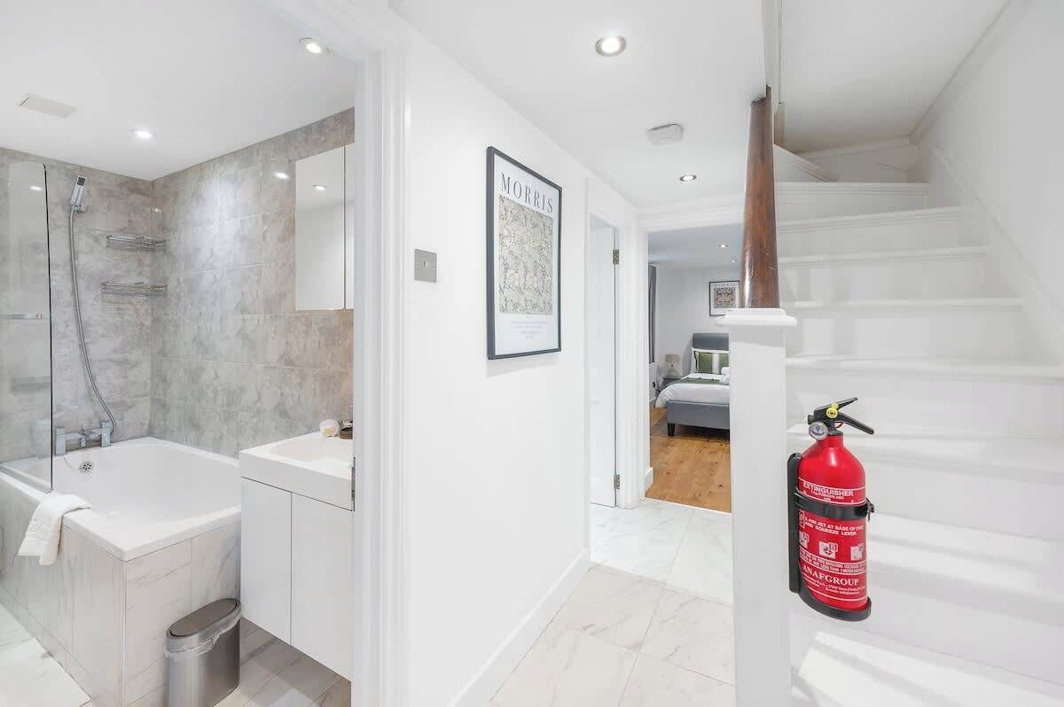 A modern bathroom with a bathtub, a shower, and a toilet. There is a framed picture on the wall and a fire extinguisher mounted on the wall.