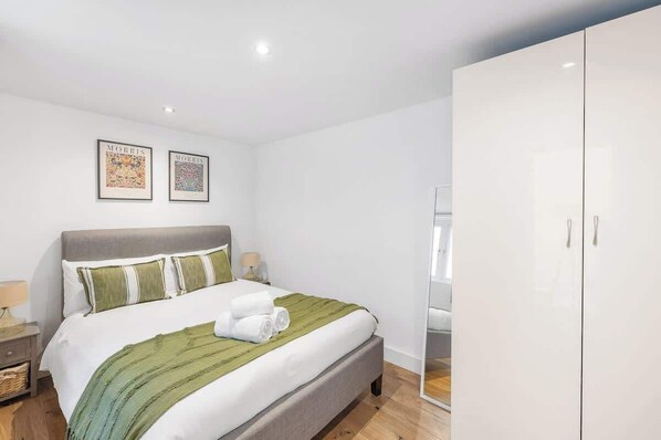Uma cama bem arrumada com um edredom verde listrado e almofadas brancas, um guarda-roupa branco e duas imagens emolduradas na parede.
