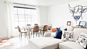 A modern living room with a sectional sofa, a dining table with chairs, a large window with curtains, and a wall-mounted artwork of a cow.