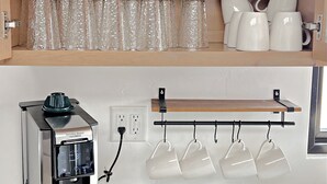A kitchen with wooden shelves holding plates, bowls, and glasses. Below, a coffee maker is on the counter.