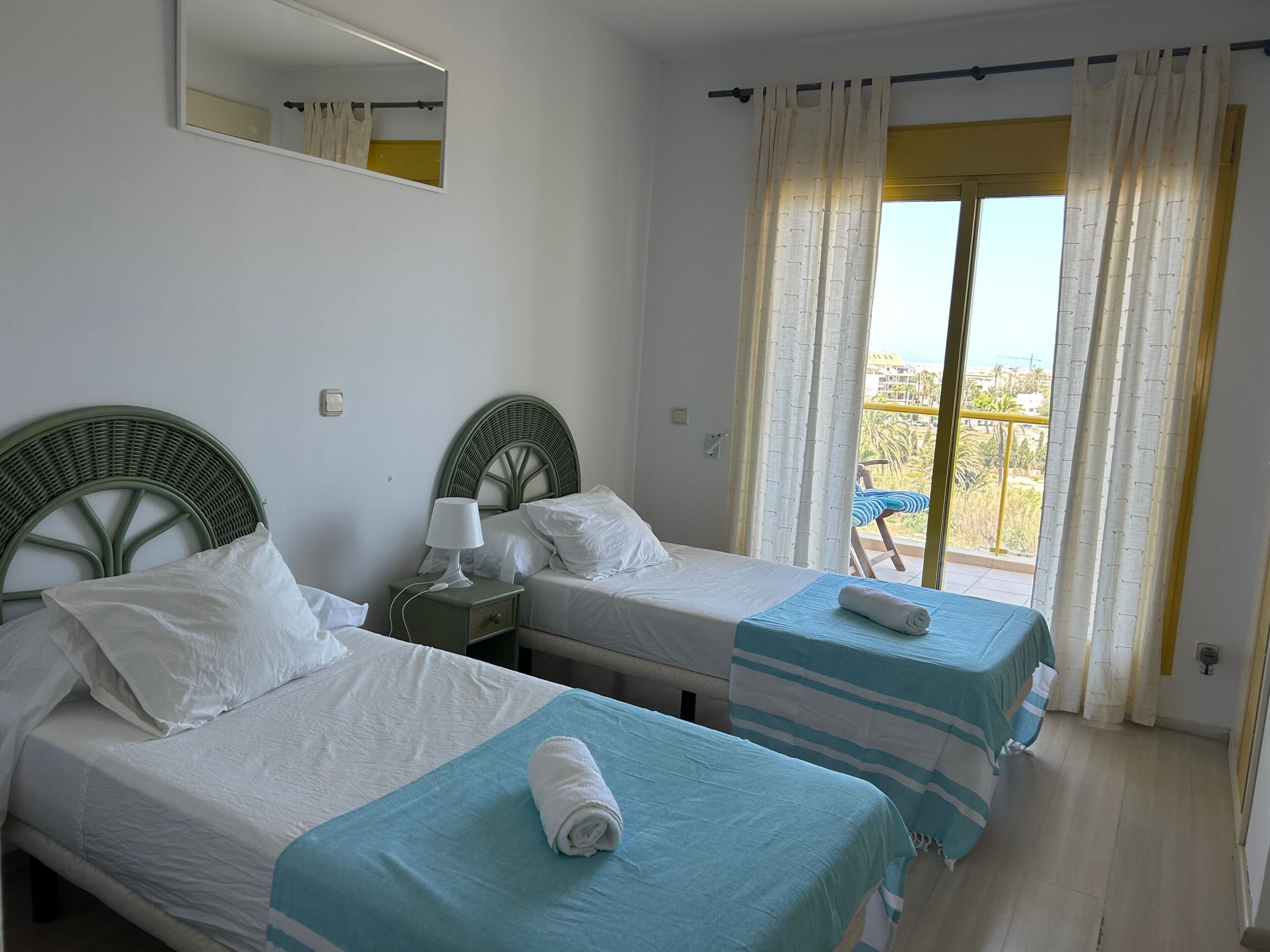 Ein Zimmer mit zwei Einzelbetten, einem Nachttisch mit Lampe, einem Spiegel und Blick ins Freie durch ein Fenster mit Vorhängen.
