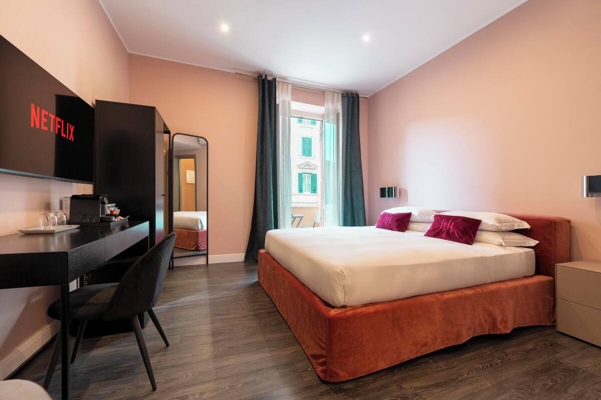 A modern hotel room with a bed, a desk with a computer, a chair, and a TV mounted on the wall.