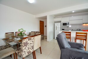 A modern kitchen with white cabinets, a microwave, and a refrigerator. There is a dining area with a glass table and patterned chairs. - Residencial João Victor - Ingleses Norte (Florianópolis)