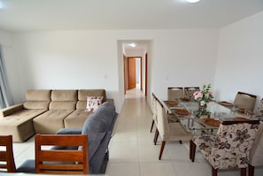 Uma sala de estar com sofá modular, uma mesa de jantar posta para quatro pessoas e um corredor que dá para outro cômodo.