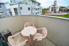 A balcony with a table and chairs, a trash bin, and a view of modern buildings in the background. - Residencial João Victor - Ingleses Norte (Florianópolis)