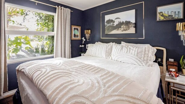 Un dormitorio con una cama grande, una ventana con cortinas, una mesita de noche con un reloj y cuadros enmarcados en la pared.