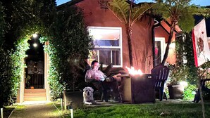 Una casa con fachada roja, una ventana y una puerta. Hay una persona sentada afuera, y un perro está acostado en el suelo.