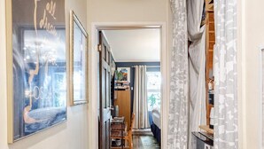 Un pasillo con un tapete estampado, un cuadro en la pared y vista a un dormitorio con cama y cortinas.