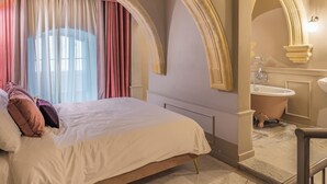 A bedroom with a bed, a window with curtains, an arched doorway, and a bathroom with a bathtub and sink. - House of W (Valletta)