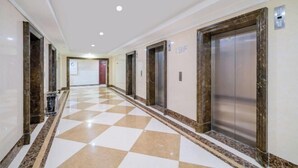 Een hotelgang met zwart-witte geruite vloer, marmeren wanden en twee liften.