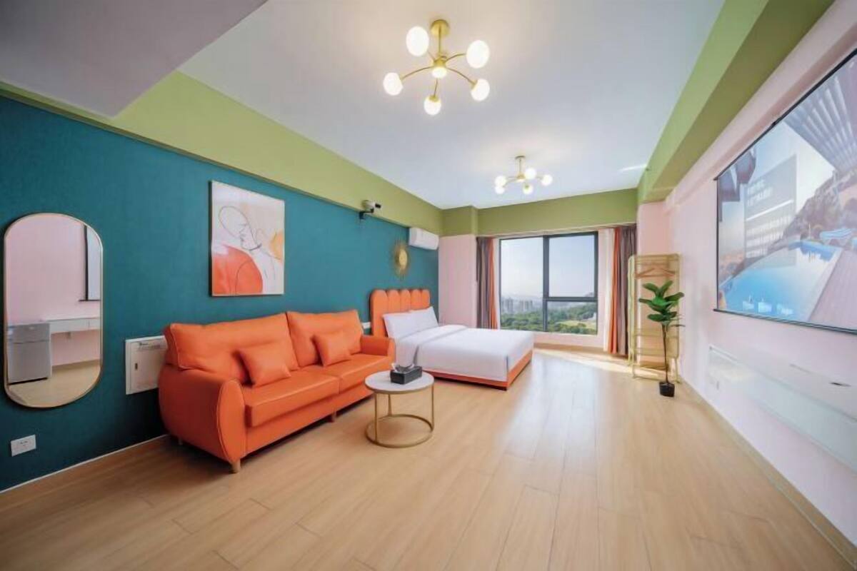 Ein modernes Hotelzimmer mit einer türkisfarbenen Akzentwand, einem großen Bett, einem orangefarbenen Sofa, einem runden Spiegel und Blick durch ein großes Fenster.