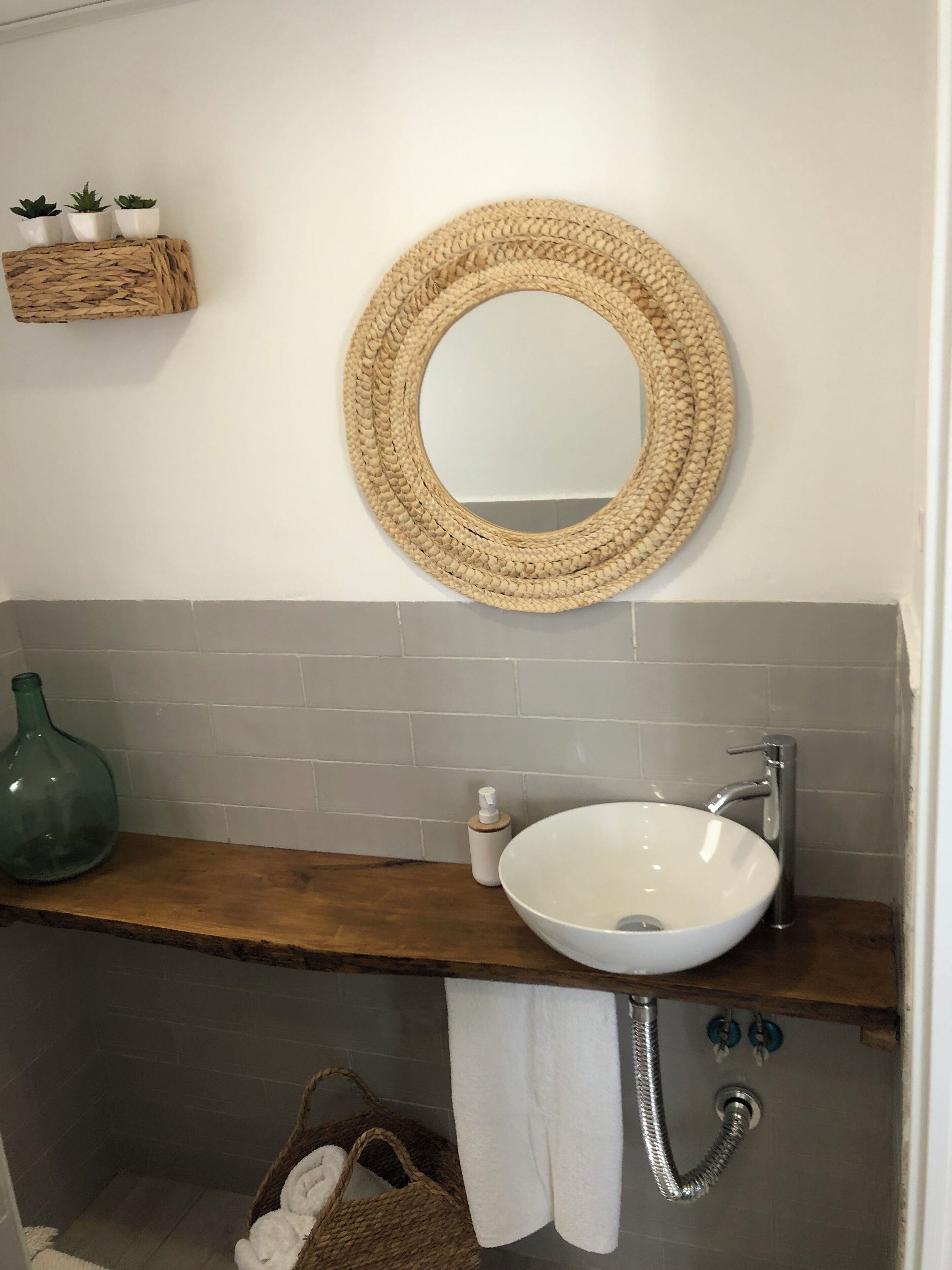 Un baño con un espejo redondo tejido, un lavabo blanco y un estante de madera.