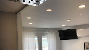 A hotel room with a large window, a flat-screen TV mounted on the wall, a wicker chair, and a bed with a patterned bedspread. - Estudios Peñiscola  (Peñíscola)