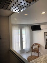 Camera d'albergo con un'ampia finestra, una TV a schermo piatto montata a parete, una poltrona in vimini e un letto con copripiumino decorato.
