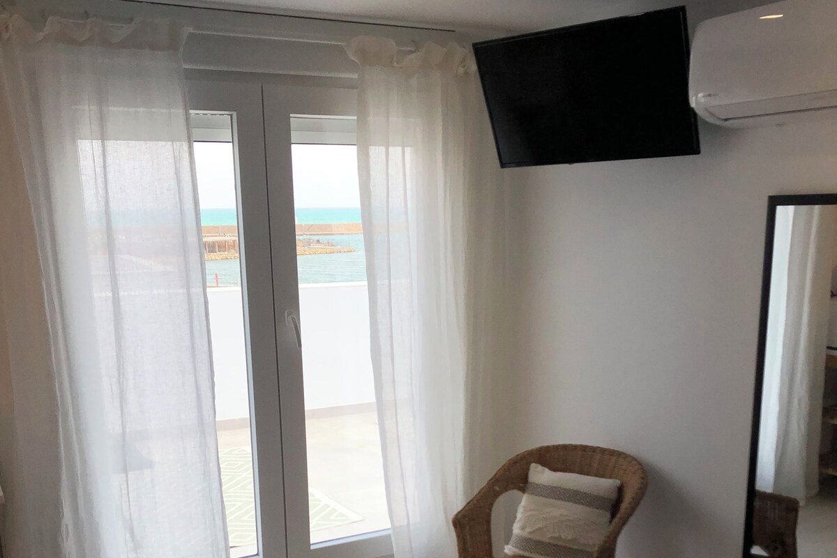 A bedroom with a bed, a chair, a large window with curtains, a flat-screen TV, and a ceiling fan.