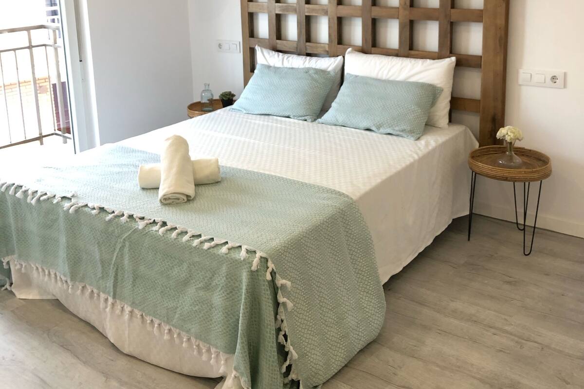 A bedroom with a wooden headboard, a bed with a green and white bedspread, a small round side table with a vase, and a balcony door.