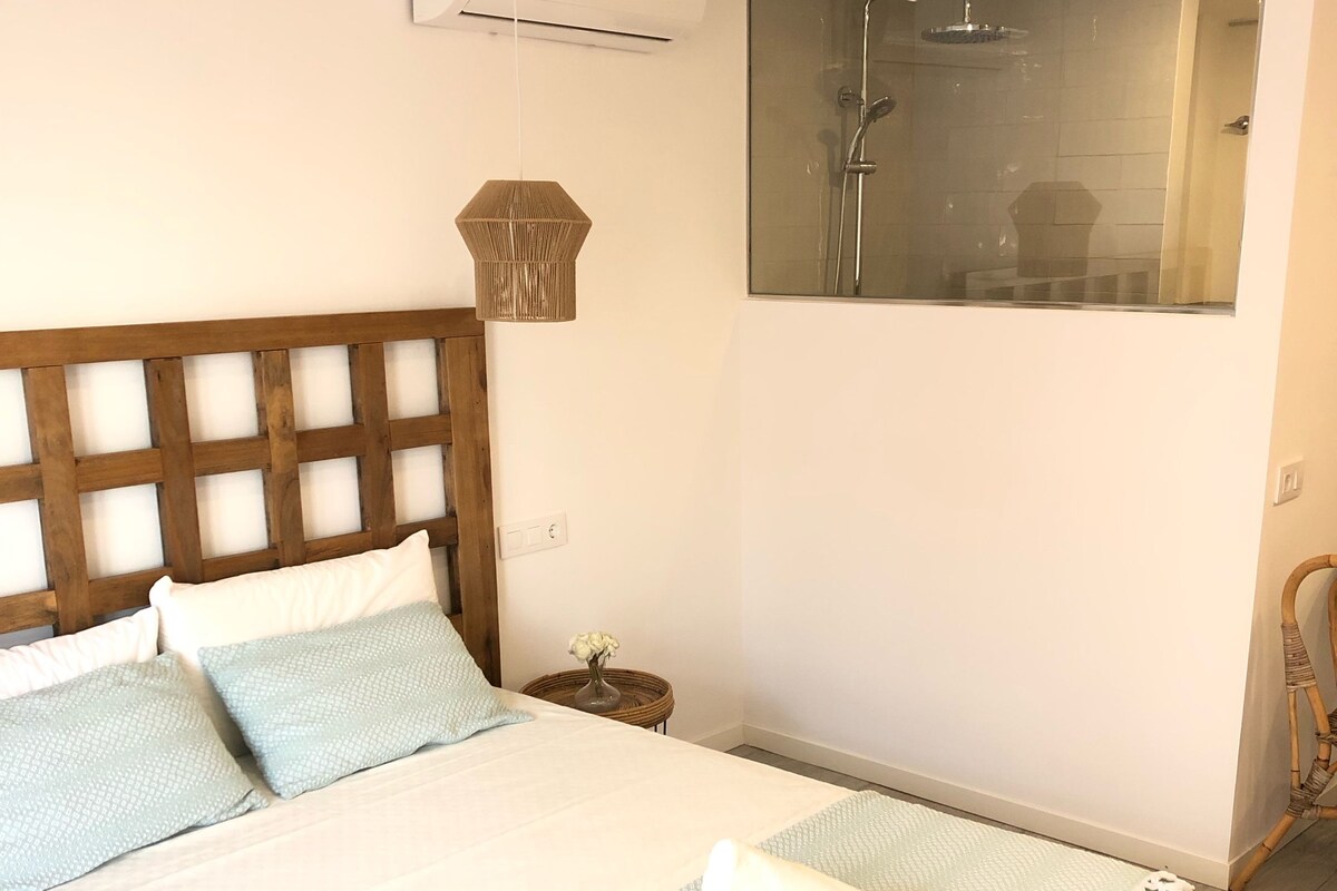 A bedroom with a wooden headboard, a bed with light blue bedding, a bedside table with a vase, and a window with a view of a shower area.