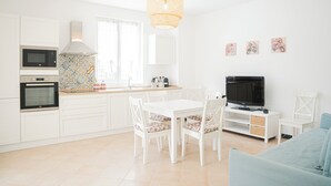 Une cuisine moderne avec des meubles blancs, un four à micro-ondes, un four et un coin repas avec des chaises et une table blanches. On y trouve un téléviseur posé sur un meuble et un luminaire suspendu en osier au-dessus de la table à manger.