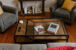A living room with a glass-top coffee table, two grey sofas, a wooden side table with books, and a remote control. - Cielo Casa on “The Hill” - Hot Tub + Spacious 3BR - Near BJC, SLU & Downtown (St. Louis)