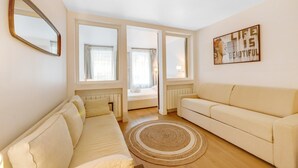 Ein modernes Wohnzimmer mit einer Couch, einem kleinen Sofa und einem großen Spiegel. Zwei Fenster mit Vorhängen, ein Heizkörper und ein Teppich sind auf dem Boden zu sehen.
