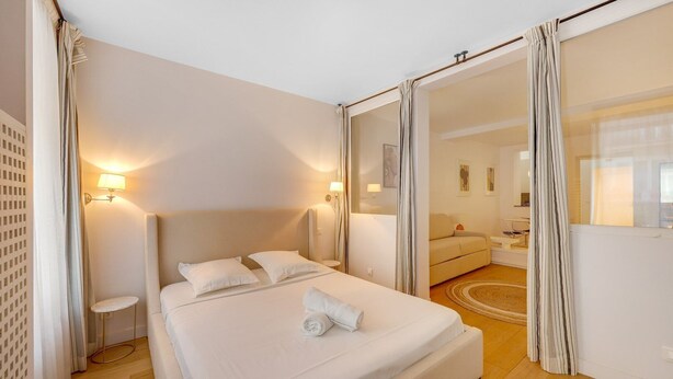 Ein Hotelzimmer mit einem großen Bett, Nachttischlampen, einem kleinen Tisch und Blick auf den Wohnbereich durch eine offene Tür.