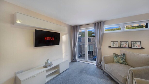 A modern living room with a flat-screen TV displaying 'NETFLIX', a white entertainment unit, a beige sofa, and a balcony with a view of buildings.