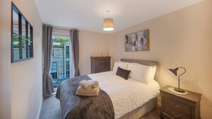 A bedroom with a bed, a nightstand, a lamp, a dresser, a window with curtains, and a painting on the wall.