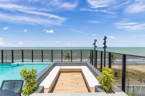 Ein Pool auf dem Dach mit Holzterrasse, umgeben von einem Glasgeländer, mit Blick auf einen Strand und das Meer.