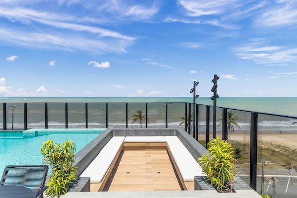 A rooftop pool with a wooden deck, surrounded by a glass balustrade, overlooking a beach and ocean. - Beach Haus by Carpediem (João Pessoa)