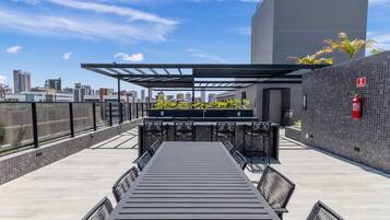 Een terras op het dak met een lange tafel en stoelen, een pergola en uitzicht op de stadshemel.