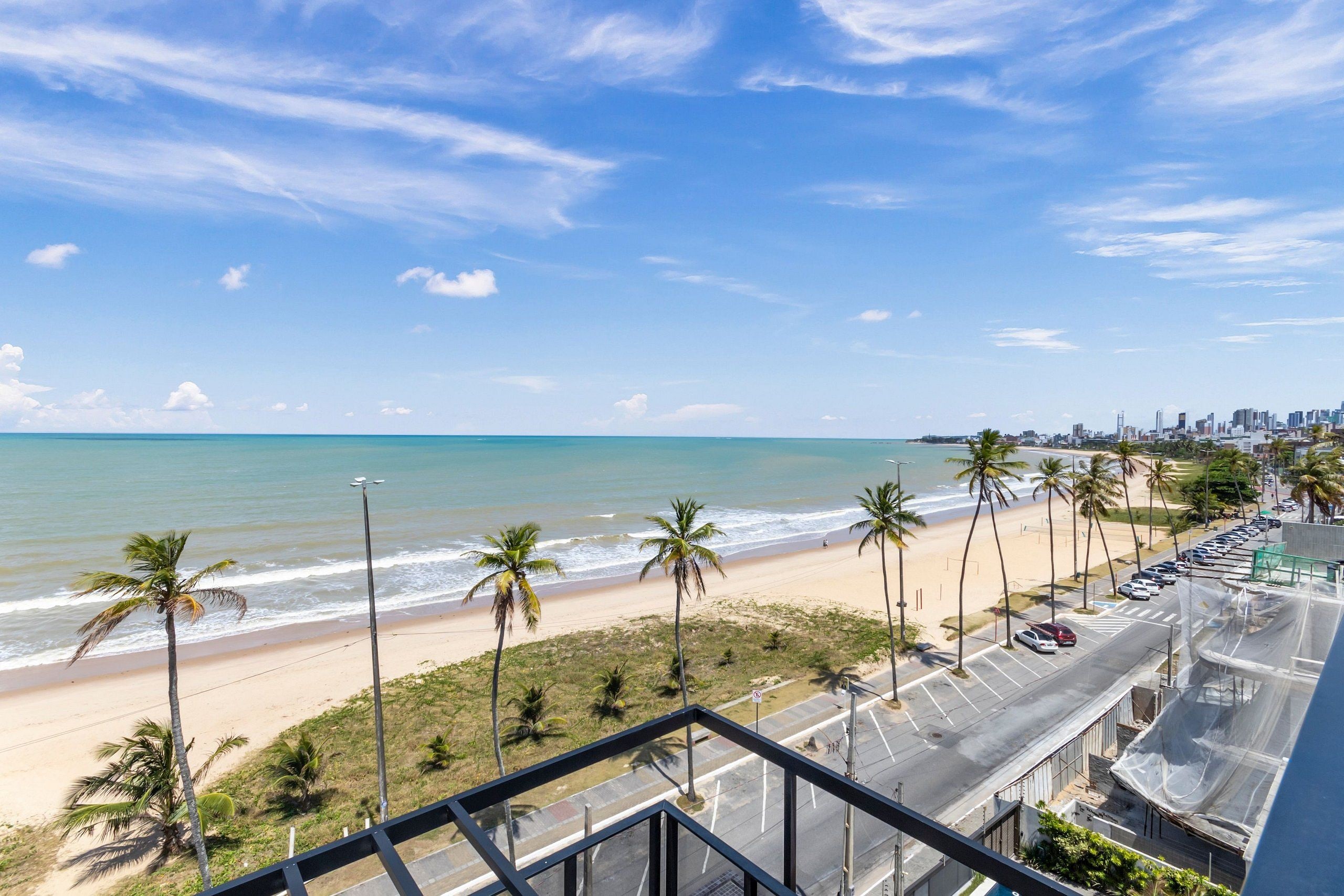Uitzicht op een strand vanaf een hoog punt, met een weg, geparkeerde auto's, palmbomen en een verre skyline van een stad.
