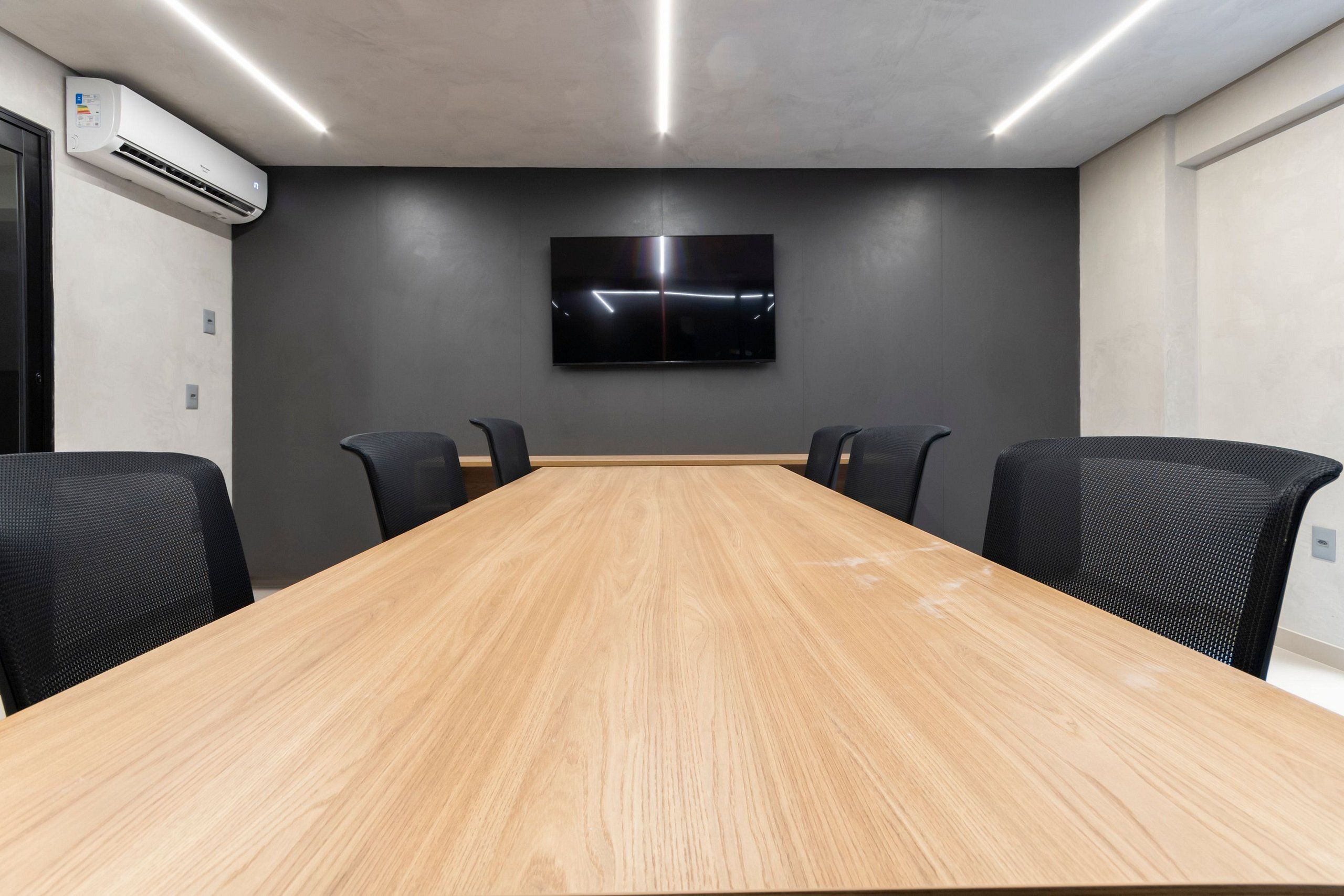 Uma sala de conferências com uma longa mesa de madeira, quatro cadeiras pretas, uma TV de tela plana na parede e um aparelho de ar condicionado à esquerda.