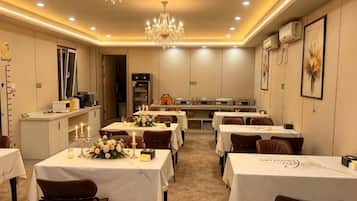 A hotel dining area with tables set for guests, a chandelier, and a kitchen in the background.