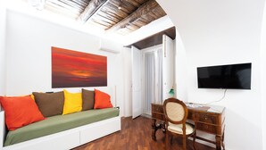 Ein Zimmer mit Holzboden, einem weißen Sofa mit bunten Kissen, einem Schreibtisch mit Stuhl, einem Fernseher und einem Bild an der Wand.