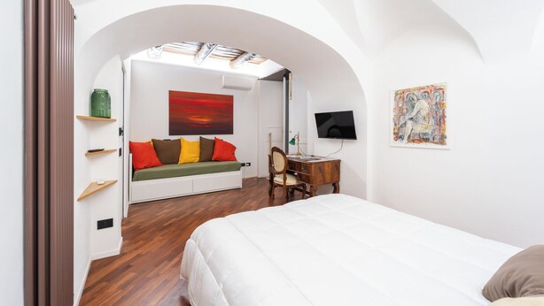Ein Schlafzimmer mit einem weißen Bett, einem Holztisch, einer grünen Vase, einem roten Gemälde und einem Fernseher an der Wand.