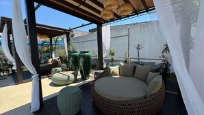 Uma área externa com assentos de vime, um banco redondo estofado e grandes vasos verdes.